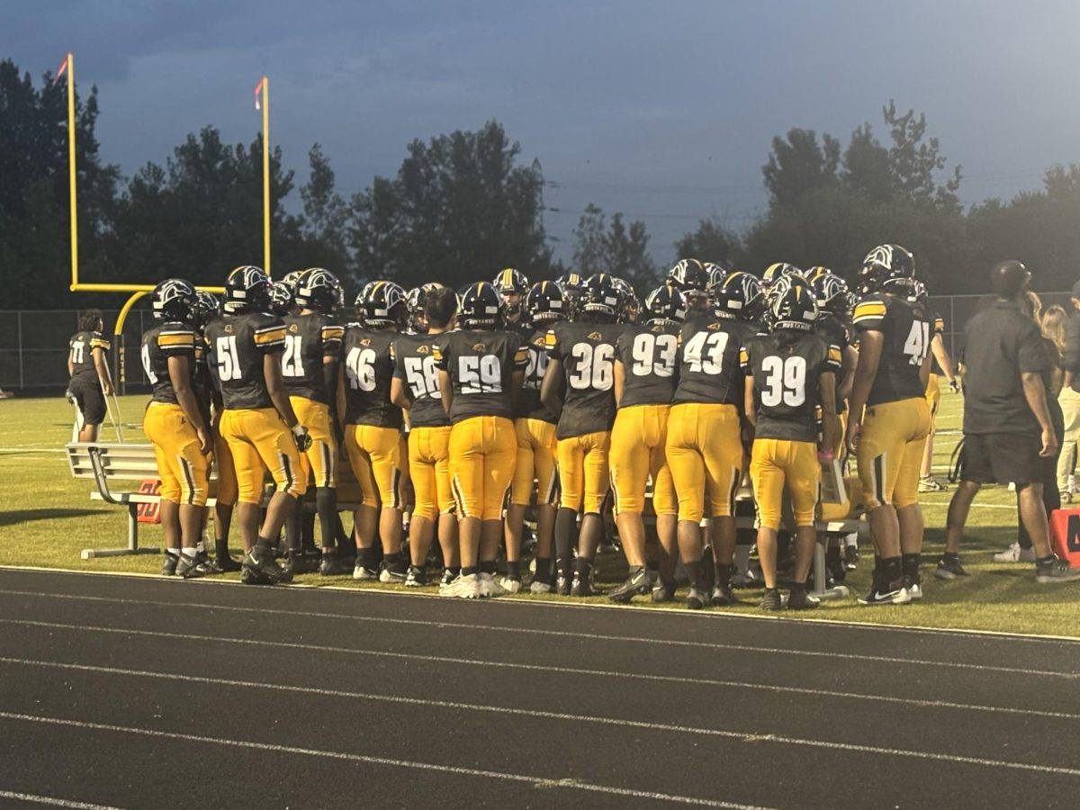 Varsity football players huddle on the side of the field, watching in support as their teammates give it their all.